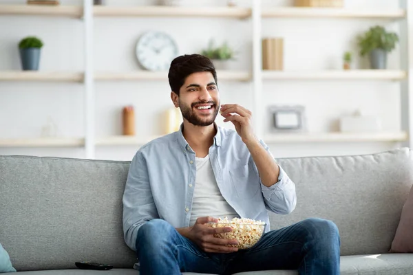 Χαλαρωμένος ανατολικός άντρας περνάει ελεύθερο χρόνο στο σπίτι, τρώει ποπ κορν και βλέπει τηλεόραση, χαλαρώνει στον καναπέ στο σαλόνι — Φωτογραφία Αρχείου