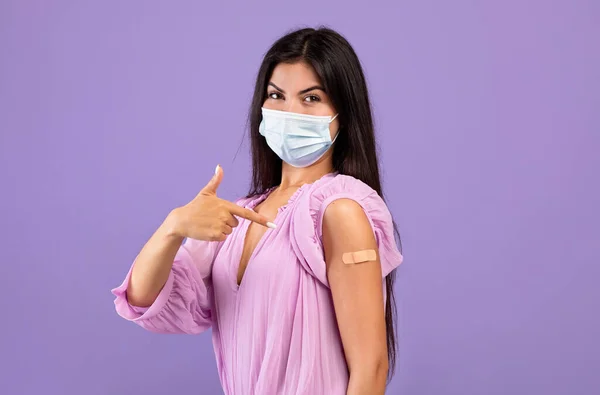 Armeense vrouw met beschermend masker wijzend vinger bij de hand met bandhulp na injectie op paarse achtergrond — Stockfoto