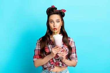 Şok olmuş, eski moda elbiseli genç bir kadın mavi stüdyo arka planında nefis milkshake içiyor.