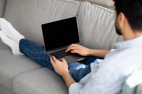 Chico árabe joven usando el ordenador portátil moderno con la pantalla negra vacía en casa, eclining en sofá, maqueta, sobre tiro del hombro — Foto de Stock