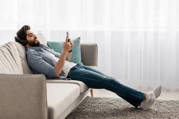 国内放松。年轻的阿拉伯男孩，带着无线耳机和智能手机，躺在舒适的沙发上，侧视 — 图库照片