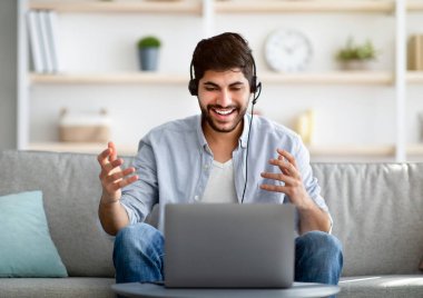 Genç Arap iş adamı dizüstü bilgisayarda kulaklık takıyor, oturma odasındaki kanepede oturuyor.