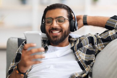 Neşeli Arap adam film izliyor, kablosuz kulaklık ve cep telefonu kullanıyor.