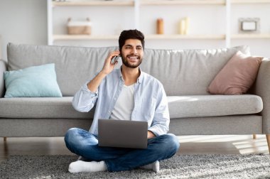Arap serbest çalışan bir adam dizüstü bilgisayar kullanıyor ve cep telefonuyla konuşuyor, oturma odasında yerde otururken çalışıyor.