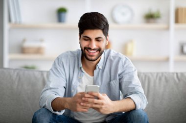 Yakışıklı gülümseyen Arap adam elinde akıllı telefonuyla oturuyor ve arkadaşlarıyla mesajlaşıyor, kanepede cep telefonuyla rahatlıyor.