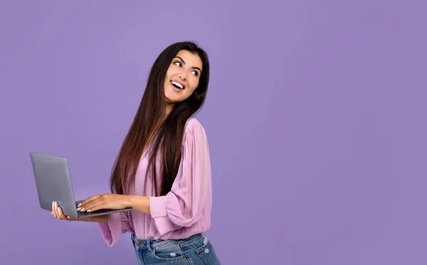 Cheerful armenian woman standing with laptop computer over purple studio background and looking back at copy space — ストック写真