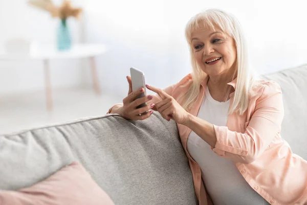 Usmívající se starší žena pomocí mobilního telefonu odpočívá na pohovce — Stock fotografie