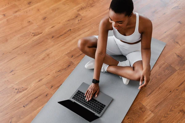 Улыбающаяся молодая черная женщина сидит на коврике для йоги с помощью ноутбука — стоковое фото