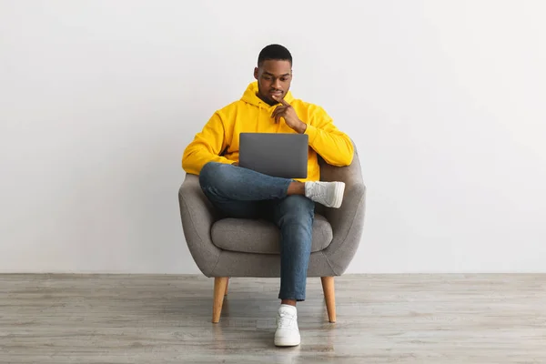 Homem preto usando laptop sentado na poltrona perto da parede cinza — Fotografia de Stock
