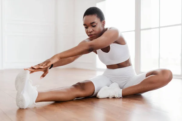 Alegre negro mujer en blanco deportivo estiramiento pierna en piso —  Fotos de Stock