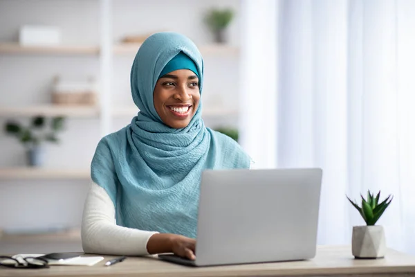 Carrière à distance. Heureuse femme musulmane noire assise au bureau avec ordinateur portable — Photo