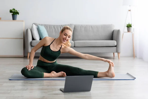 Young blonde sports woman in sportswear doing workout on floor at home and watching fitness videos in laptop — Stock Photo, Image