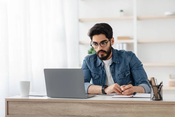 Homme arabe concentré prenant des notes et regardant l'ordinateur portable, écrivant des idées de projet dans un cahier assis sur le lieu de travail — Photo