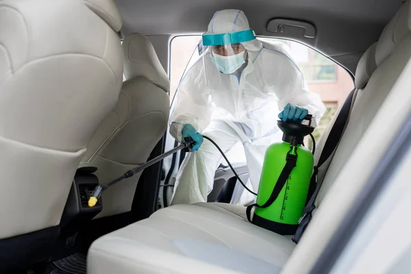 Hombre en traje de materiales peligrosos con máscara desinfectando el interior del coche, limpie las superficies limpias que se tocan con frecuencia — Foto de Stock