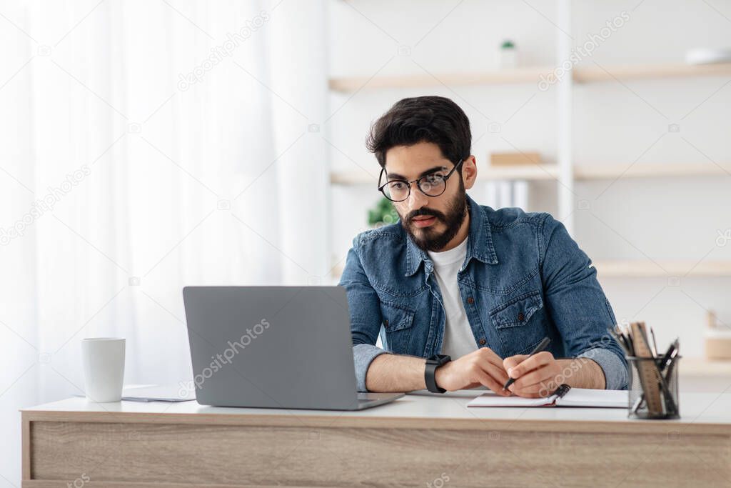 Concentrated arab man taking notes and looking at laptop, writing project ideas in notebook sitting at workplace