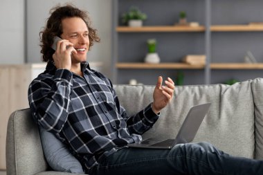 Evde dizüstü bilgisayar kullanarak cep telefonuyla konuşan mutlu serbest çalışan erkek.
