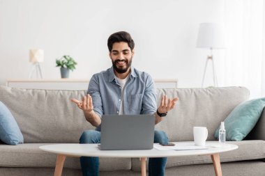 Genç Arap serbest yazar dizüstü bilgisayarla video görüşmesi yapıyor, evdeki kanepede otururken internet konferansı yapıyor, boş alan.