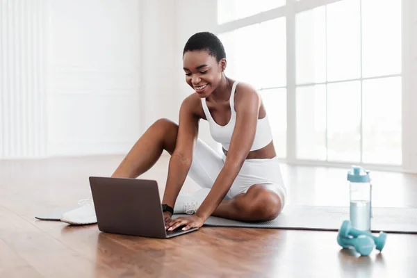 Усміхнена молода чорна жінка сидить на килимку йоги за допомогою ноутбука — стокове фото