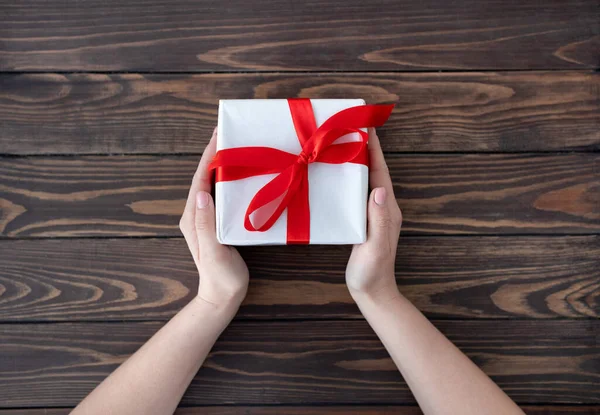 Vrouw geven gewikkelde kerst of andere vakantie met de hand gemaakt in wit papier met rood lint, houten tafel, bovenaanzicht — Stockfoto
