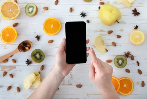 Dieting concept. Lady holding cellphone with blank black screen on wooden table with fruit slices, top view, mockup — Stock Photo, Image