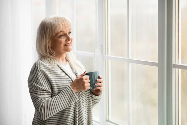Зрелая женщина пьет горячий кофе, глядя в окно — стоковое фото