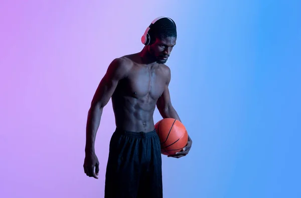 Porträt eines motivierten schwarzen Basketballers mit nacktem Oberkörper, der Kopfhörer trägt und Ball im Neonlicht hält — Stockfoto