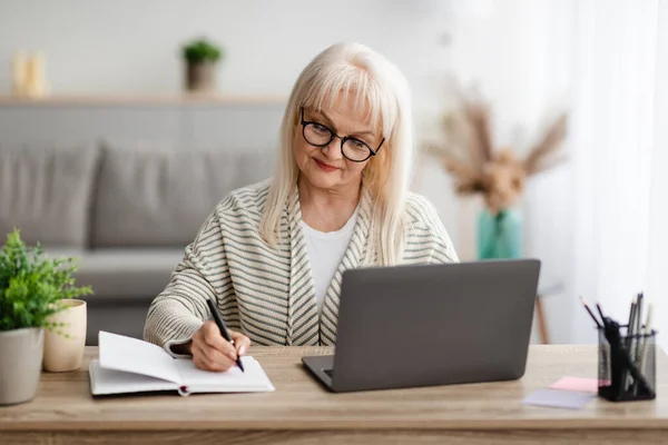 Mature lady writing in notebook and using laptop at home — Stock Photo, Image