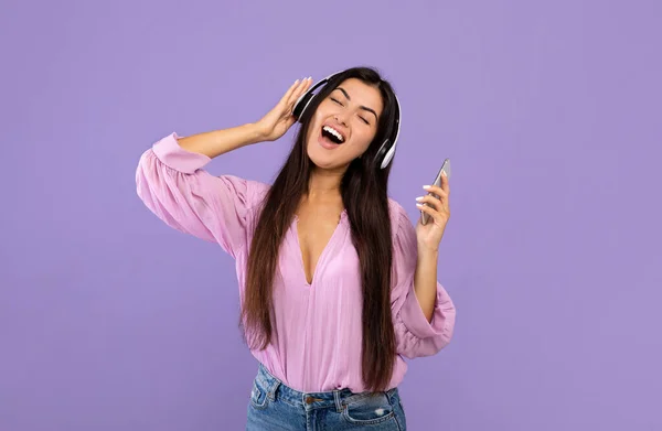 Bonne dame arménienne avec smartphone et écouteurs sans fil, écouter de la musique et chanter sur fond violet — Photo