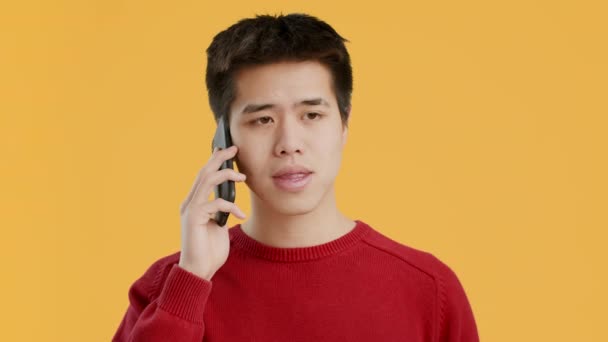 Азиатский парень разговаривает по мобильному телефону, общаясь на фоне жёлтой студии — стоковое видео