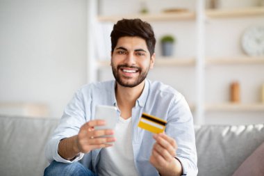 İnternetten mal sipariş ediyorum. Evde internet üzerinden kredi kartı ve akıllı telefon alışverişi yapan mutlu genç bir Arap.