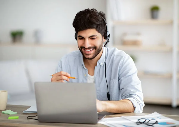 Homem árabe animado em fones de ouvido estudando on-line com laptop em casa, assistindo webinar e tomar notas para notepad — Fotografia de Stock