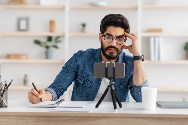 Webinárium koncepció. Koncentrált arab fickó szemüveggel jegyzetel és online előadást néz okostelefonon, másolja a helyet — Stock Fotó