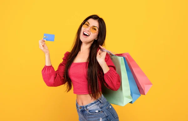 Lätt att handla. Överlycklig armeniska kvinna som innehar kreditkort och ljusa shopper påsar, poserar över gul bakgrund — Stockfoto
