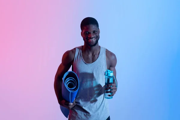 Yoga para uma vida saudável. Jovem negro feliz segurando tapete esportivo e garrafa de água em luz de néon — Fotografia de Stock