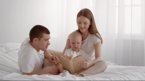 Šťastní mladí rodiče čtení knihy na jejich roztomilé dítě doma — Stock video