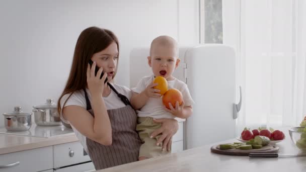 Az anyaság kihívásai. Elfoglalt nő a kezében a baba és beszél a mobilon a konyhában — Stock videók