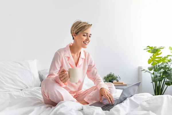 Ευτυχισμένη γυναίκα που κάθεται στο κρεβάτι χρησιμοποιώντας υπολογιστή πίνοντας καφέ — Φωτογραφία Αρχείου