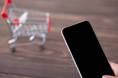 Ahşap masada içi boş ekranlı akıllı telefonu olan ve arka planda mini alışveriş arabası olan bir kadın.