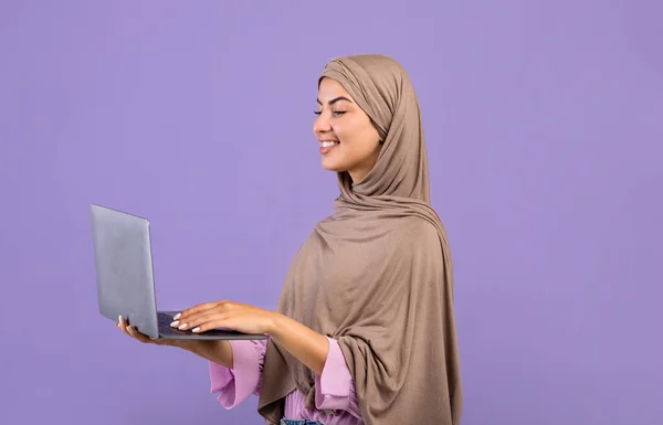 Mujer musulmana feliz con el ordenador portátil en las manos sobre fondo de estudio púrpura, sonriente dama islámica en hijab sosteniendo ordenador PC —  Fotos de Stock