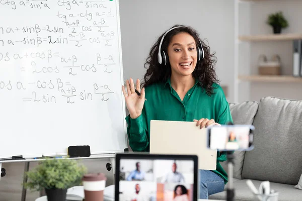 Smiling young woman teaching maths to students online, waving hello