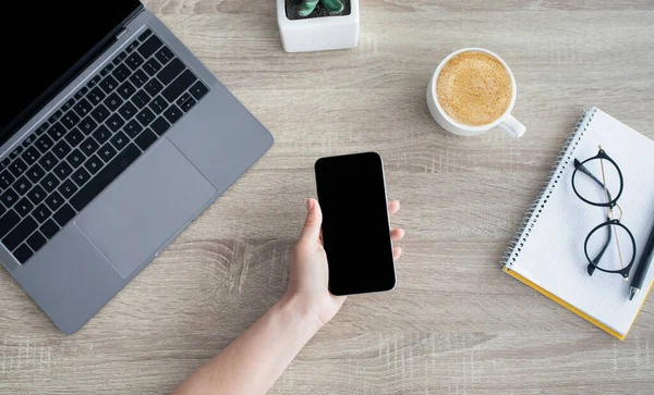 Tangan wanita memegang ponsel dengan layar hitam kosong untuk iklan Anda, duduk di tempat kerja dengan laptop, tampilan atas — Stok Foto