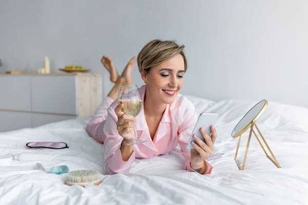 Lycklig kvinna liggande i sängen läser textmeddelande dricka alkohol — Stockfoto