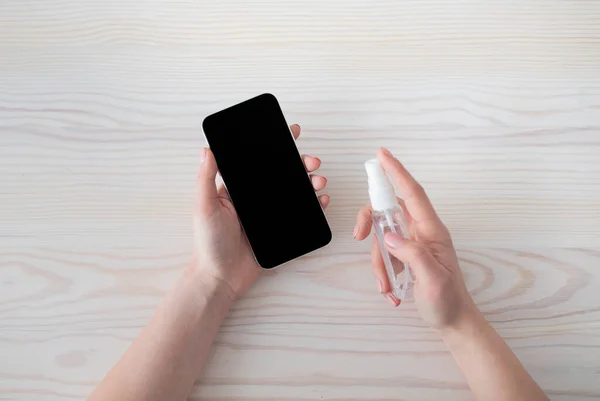 Gadget disinfection. Unrecognizable woman sanitizing smartphone with disinfectant mist spray, top view — Stock Photo, Image