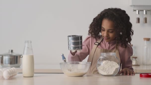 Adolescente afroamericana che si prepara la pasticceria a casa, aggiunge farina con setaccio metallico all'impasto, cucina in cucina — Video Stock