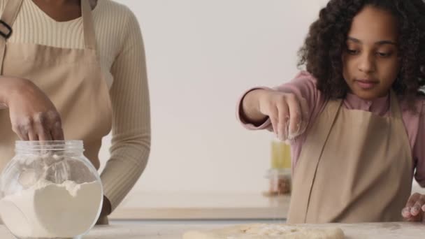 Piccola ragazza afroamericana che studia per cuocere con sua madre, fiorendo pasta cruda sul tavolo prima di rotolare fuori — Video Stock