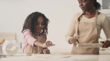 Küçük Afrikalı Amerikalı kızın portresini büyüt. Annesine mutfakta yemek yaparken yardım et, kadın hamur toplarken.