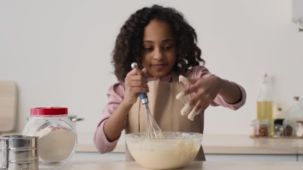 Kleine afrikanische Amerikanerin bereitet Teig in der Küche zu, macht sich die Finger mit süßem Gebäck schmutzig und leckt sie — Stockvideo