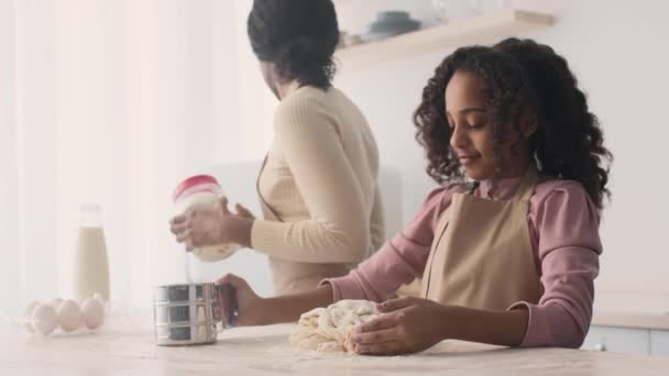 Yoğunlaşmış küçük Afrikalı Amerikalı kız un sürüyor ve hamur yoğuruyor, annesi mutfaktaki malzemeleri çıkarıyor. — Stok video