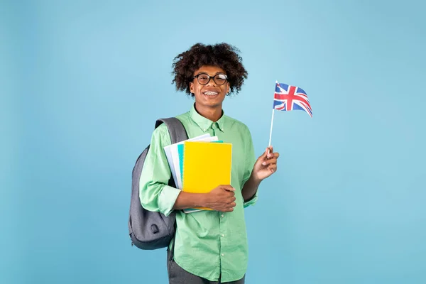 Šťastný africký americký dospívající výměnný student s notebooky drží britskou vlajku, usmívá se na kameru na modrém pozadí studia — Stock fotografie