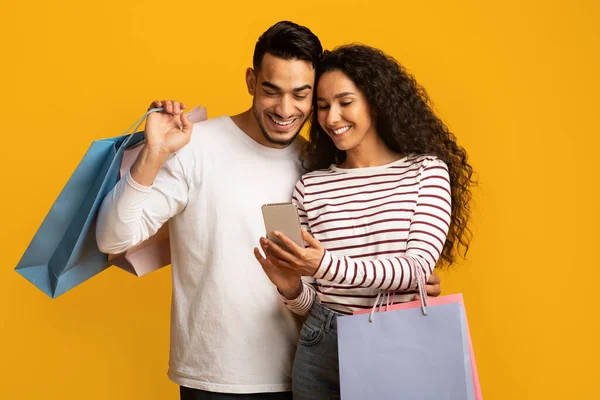 Aplicación de descuento. Alegre pareja árabe sosteniendo Smartphone y coloridas bolsas de compras de papel —  Fotos de Stock